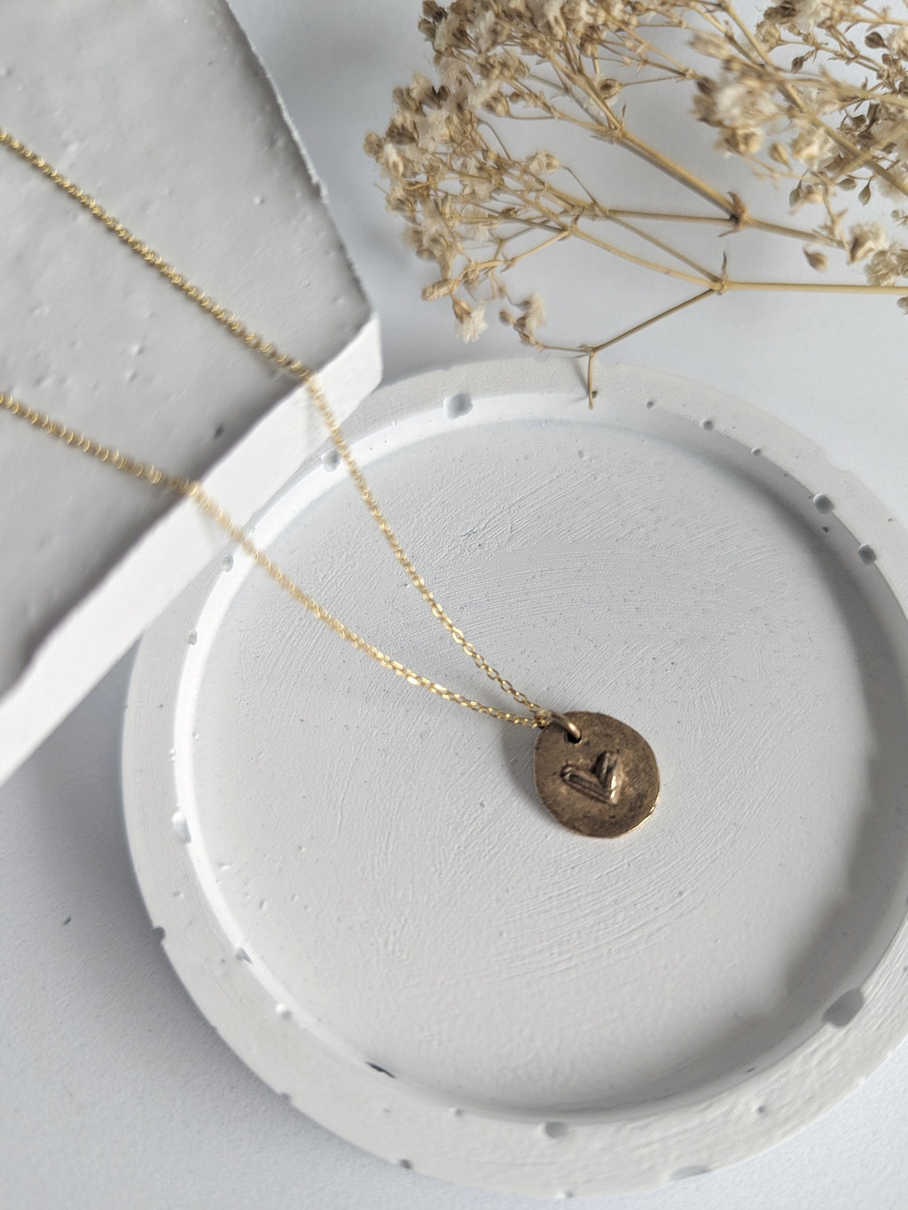 Gold necklace with a round pendant on a white dish, with dried flowers in the background.