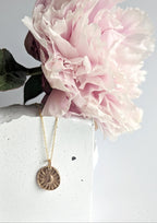 Gold necklace with a circular pendant displayed against a pink flower on a white surface.