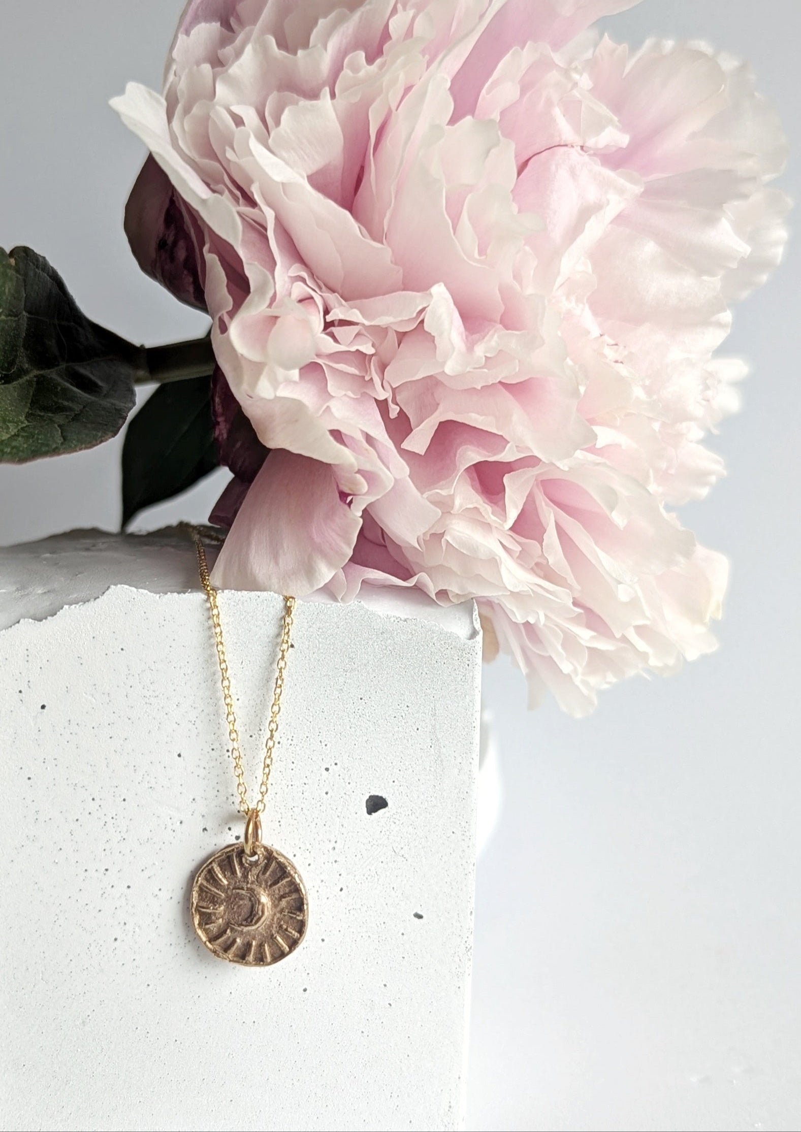 Gold necklace with a circular pendant displayed against a pink flower on a white surface.